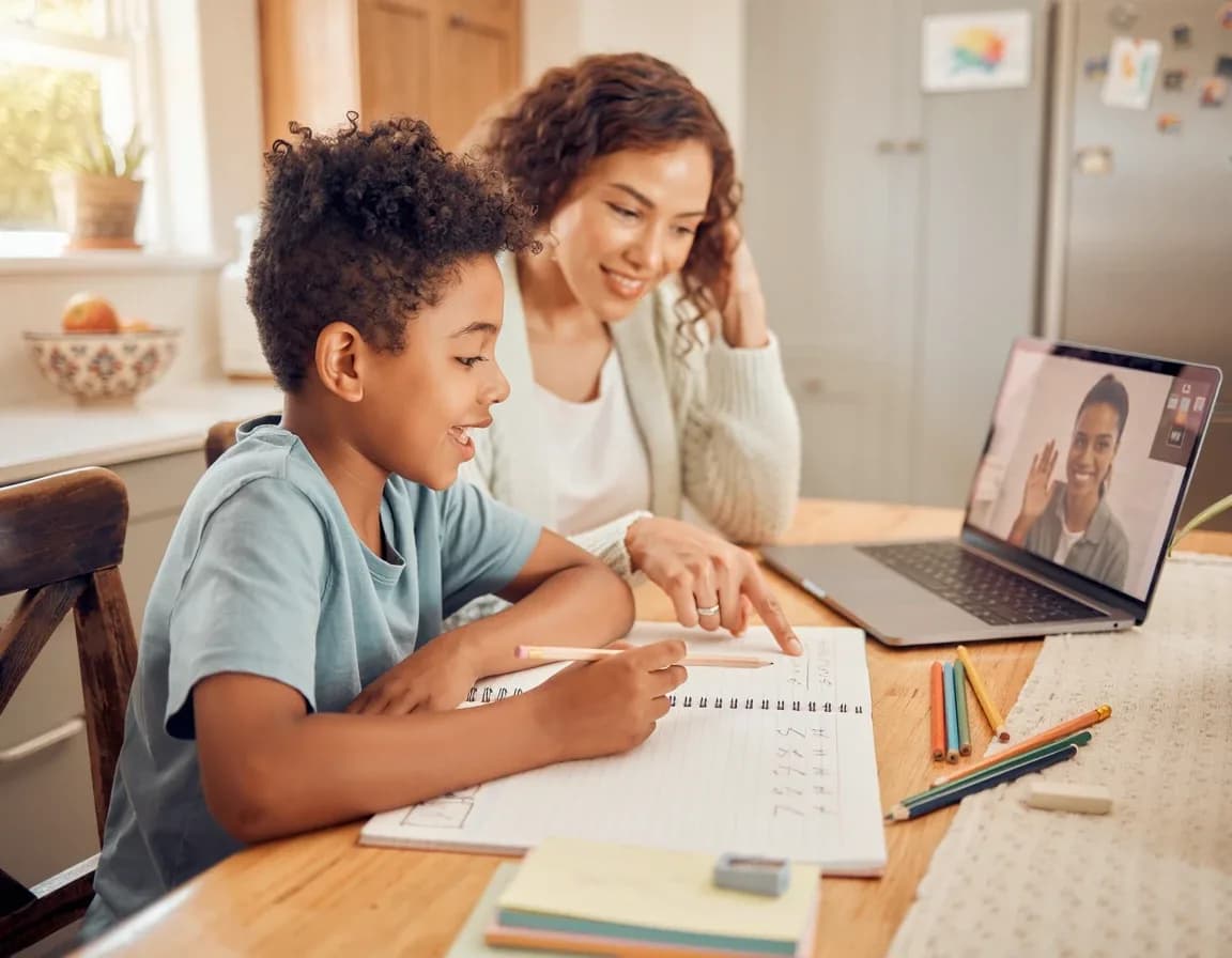 Un enfant fait ses devoirs à la table de la cuisine, accompagné par un adulte, devant un ordinateur portable où apparaît son enseignante.